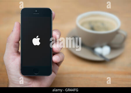 Un homme se penche sur son iPhone qui affiche le logo Apple sur un fond noir, avec une tasse de café (usage éditorial uniquement). Banque D'Images