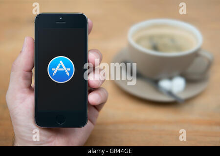 Un homme se penche sur son iPhone qui affiche le logo Apple App Store, tandis que sam avec une tasse de café (usage éditorial uniquement). Banque D'Images