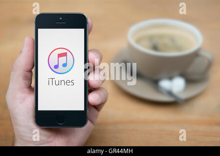 Un homme se penche sur son iPhone qui affiche le logo Apple iTunes, alors qu'assis avec une tasse de café (usage éditorial uniquement). Banque D'Images