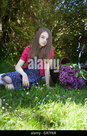 Teenage girl sitting dans un champ avec un vélo avec un panier de fleurs en arrière-plan à tout droit à l'appareil photo Banque D'Images