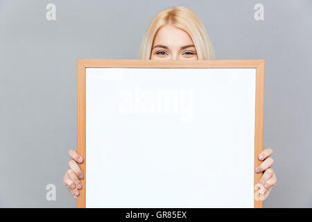 Cute blonde jeune femme cachant son visage derrière un tableau blanc en blanc sur fond gris Banque D'Images