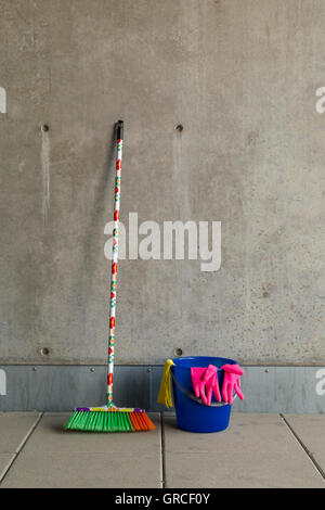 L'équipement de nettoyage colorés (balai, seau, chiffon et gants) en face d'un mur de béton gris, monotone et plancher teint. Banque D'Images