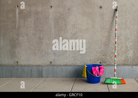 L'équipement de nettoyage colorés (balai, seau, chiffon et gants) en face d'un mur de béton gris, monotone et plancher teint. Banque D'Images