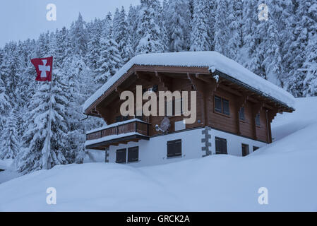 Swiss Chalet en face de la forêt de sapins couverts de neige profondément avec de brandir le drapeau suisse dans le vent. Dans l'avant-plan Snowslope. Banque D'Images