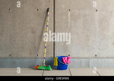 L'équipement de nettoyage colorés (balai, seau, chiffon et gants) en face d'un mur de béton gris, monotone et plancher teint. Banque D'Images