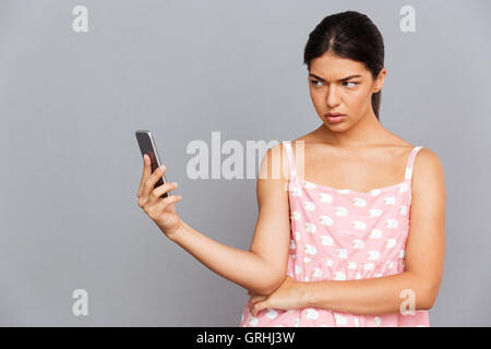 Portrait d'une femme brunette mécontent en robe rose en photo sur smartphone selfies isolé sur fond gris Banque D'Images