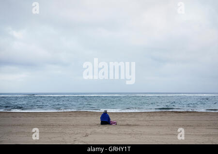 Vue arrière d'une femme âgée assise sur la plage vide. Banque D'Images