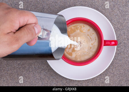 Close up de la main d'un homme verser du lait dans du café noir à partir d'une verseuse en métal alors qu'il se prépare une tasse de début de matinée énergisante Banque D'Images