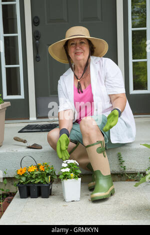 Vieille dame s'apprête à transplanter les semis de fleurs assis sur les marches de son porche dans un chapeau, gants et bottes wit Banque D'Images