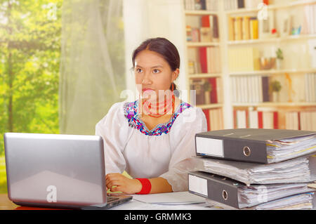 Jeune jolie fille portant des vêtements traditionnelle des Andes, assis par office de bureau avec un ordinateur portable, les reliures à anneaux empilés sur table, arrière-plan de la fenêtre de jardin Banque D'Images