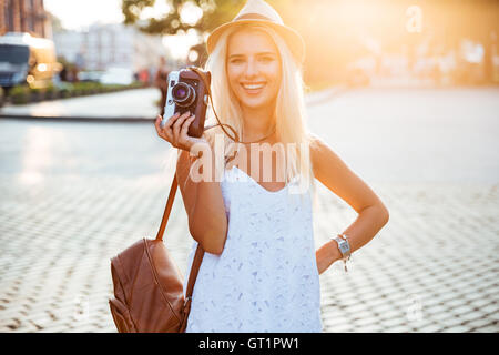 Young smiling blonde girl in hat holding caméra rétro tout en se tenant sur la rue Banque D'Images