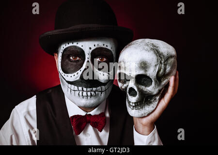 Portrait d'un jeune homme avec un mexicain comté maquillage, portant chapeau et chapeau melon, tenant un crâne dans sa main à côté de sa Banque D'Images