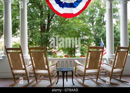 Quatre chaises à bascule en bois vacants alignés sur un patio donnant sur un jardin luxuriant en dessous d'un drapé drapeau américain symbolisant 4e Mar Banque D'Images