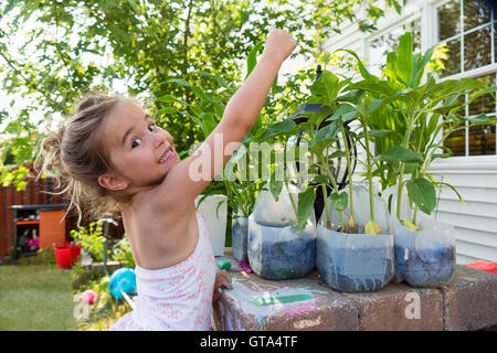 Cute little girl comparer sa taille à la plantation de fleurs qu'elle est réutilisée dans la bouteille de lait en plastique à l'extérieur près de house Banque D'Images