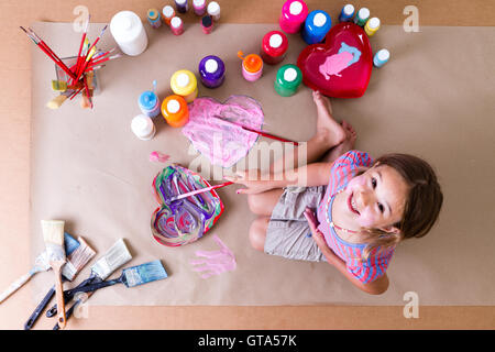 Jolie petite fille agenouillée artiste créative sur papier brun avec ses pinceaux peintures et dessins colorés et de coeur Banque D'Images