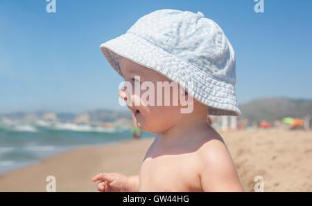 Happy Baby Boy with blue hat assis sur le sable et l'observation de la mer sur la plage Banque D'Images