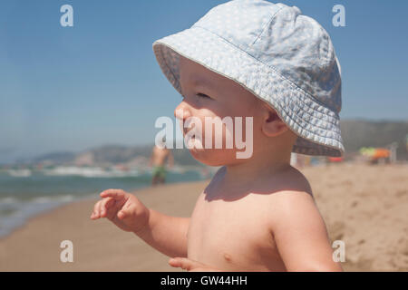 Happy Baby Boy with blue hat assis sur le sable et l'observation de la mer sur la plage Banque D'Images