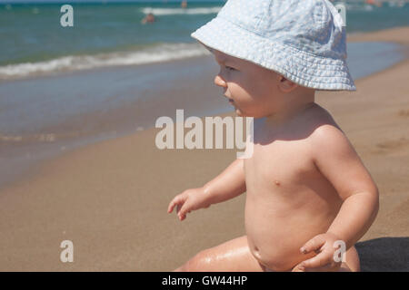 Happy Baby Boy with blue hat assis sur le sable et l'observation de la mer sur la plage Banque D'Images