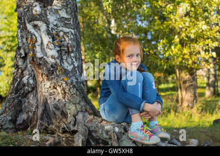 Petite fille assise sur les racines d'un bouleau. Banque D'Images