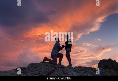 Paysage avec des silhouettes d'un homme faire de demande en mariage à sa petite amie sur le sommet de la montagne au soleil colorés. Banque D'Images