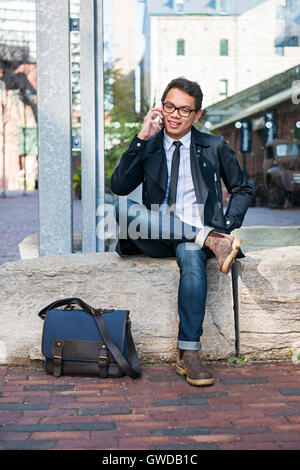 Young Asian business man talking on mobile phone assis dehors on city street Banque D'Images