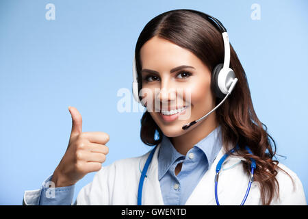 Portrait of happy smiling young doctor in casque, showing thumb up sign geste, sur fond bleu lumineux Banque D'Images