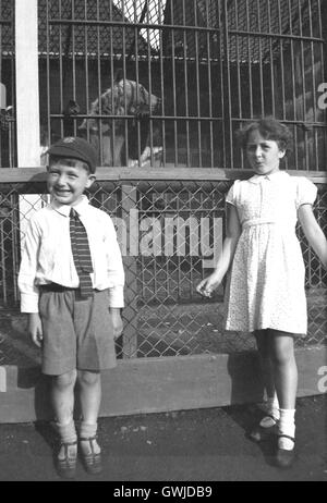 Années 1930, historiques, frère et sœur dans l'uniforme scolaire à un zoo, posant pour une photo en face de l'ours dans leur cage. Banque D'Images