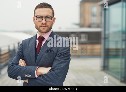 Confiant classy businessman wearing glasses debout sur un balcon en plein air à la recherche de l'appareil photo avec soin le bras plié Banque D'Images