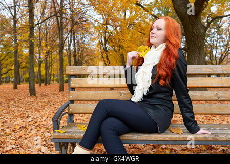 Belle jeune fille portrait s'asseoir sur le banc de parc avec feuille jaune dans la main, automne, rousse, cheveux longs Banque D'Images