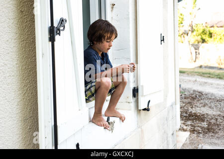 Garçon assis sur le rebord de la maison de vacances à regarder le bracelet, France Banque D'Images