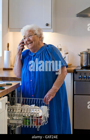 96 ans indépendant happy vieille dame âgée répond au téléphone alors qu'elle charge le lave-vaisselle dans sa cuisine maison communautaire moderne Banque D'Images