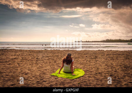 Femme assise sur la plage sur une serviette de plage vert contemplant à la mer et l'horizon devant elle. Banque D'Images
