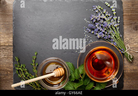 Tisane dans une tasse en verre, miel, différents types d'herbes : thym, menthe, lavande sur fond noir en noir (vue de dessus) Banque D'Images