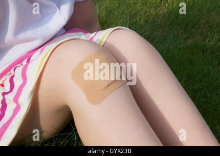 Une fille assise sur l'herbe dans une jupe à rayures avec un gros plâtre sur son genou. Banque D'Images