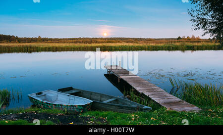 Les planches de bois jetée sur l'eau calme du lac, rivière et l'aviron deux bateaux de pêche. Lune se lever sur l'eau du soir. Banque D'Images