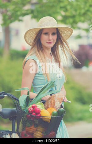 Attractive blonde woman with straw hat standing dans le parc et posant à côté de vélo avec panier plein de provisions. Processus post Banque D'Images