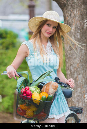 Attractive blonde womanwith straw hat debout dans le parc et posant à côté de vélo avec panier plein de provisions. Banque D'Images