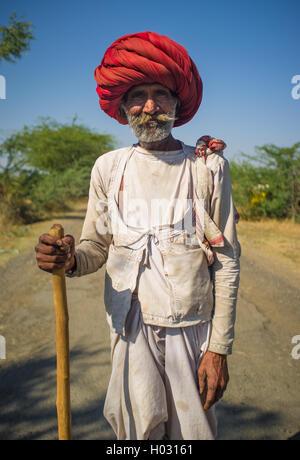 Région GODWAR, INDE - 14 février 2015 : personnes âgées Rabari tribesman avec big red turban et cane se tient sur la route. Rabari ou Rewari Banque D'Images