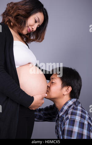 Jeune homme embrassant son ventre de femme enceinte sur fond de mur gris Banque D'Images