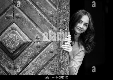 Jolie jeune fille de peering derrière l'ancienne porte en bois, en noir et blanc. Banque D'Images