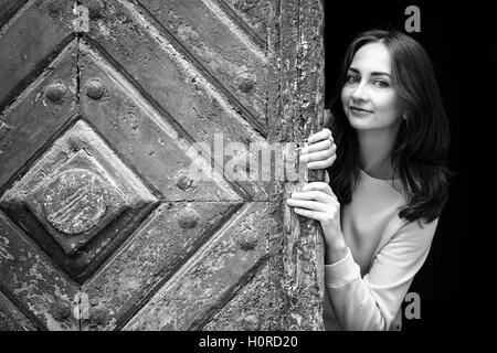 Jolie jeune fille de peering derrière l'ancienne porte en bois, en noir et blanc. Banque D'Images