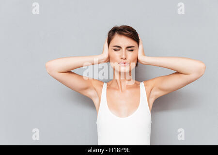 Jeune femme avec les yeux fermés couvrant ses oreilles avec des paumes isolé sur fond gris Banque D'Images