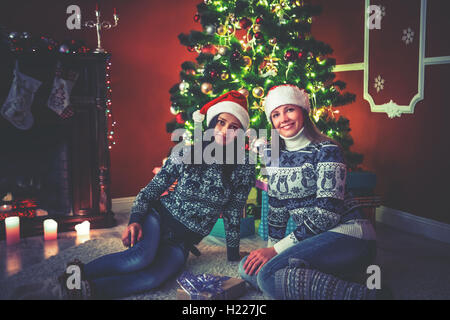 Young female friends dans chapeaux père noël près d'un arbre de Noël dans la salle décorée. Banque D'Images