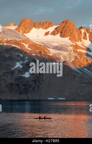 Les kayakistes pagayant en kayak double au-dessous de la montagne au coucher du soleil, l'île d'Ammassalik, Fjord Sammileq, dans l'Est du Groenland Banque D'Images