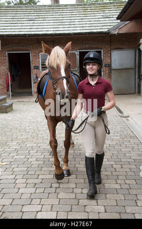 La préparation d'un jeune cavalier poney pour sa leçon. Teenage girl walking un poney dans la cour d'un stable riding school Banque D'Images