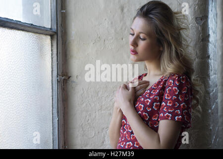 Belle jeune femme en robe rouge par la fenêtre les mains sur coeur Banque D'Images