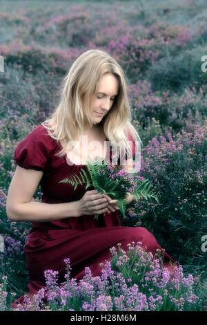 Une femme en robe rouge est assise dans la bruyère avec un bouquet de fleurs Banque D'Images