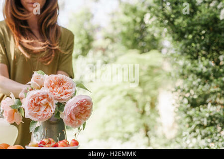 Femme debout à une table dans un jardin, d'un vase de roses et un bol de cerises. Banque D'Images