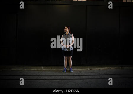 Une femme dans un T-shirt et pantalon avec des chaussures de course, debout dans l'ombre à l'extérieur d'un bâtiment sur une rue. Banque D'Images