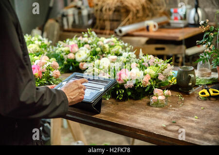 Une femme dans un atelier du fleuriste à l'aide d'une tablette numérique, à un établi avec des arrangements floraux. Banque D'Images